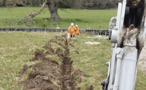 Two operatives walking up a field beside a trench they have dug out with a digger ready for installation of newt fencing