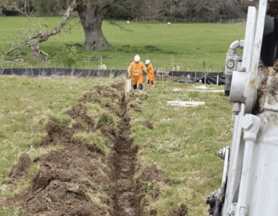 Two operatives walking up a field beside a trench they have dug out with a digger ready for installation of newt fencing